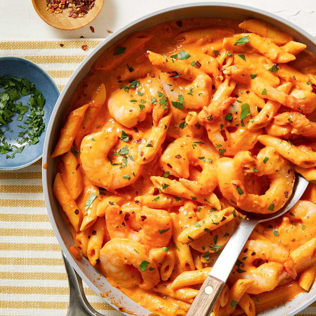 Top view shot of Creamy Tomato Shrimp with Penne; in a skillet with a serving spoon; over a napkin; sprinkle with parsley and chilli flakes; all set on a white surface