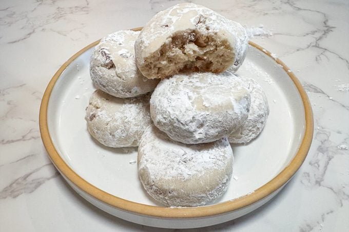 A plate of round cookies coated in powdered sugar, with one cookie broken in half to reveal a nutty filling inside. The plate sits on a light marble surface.