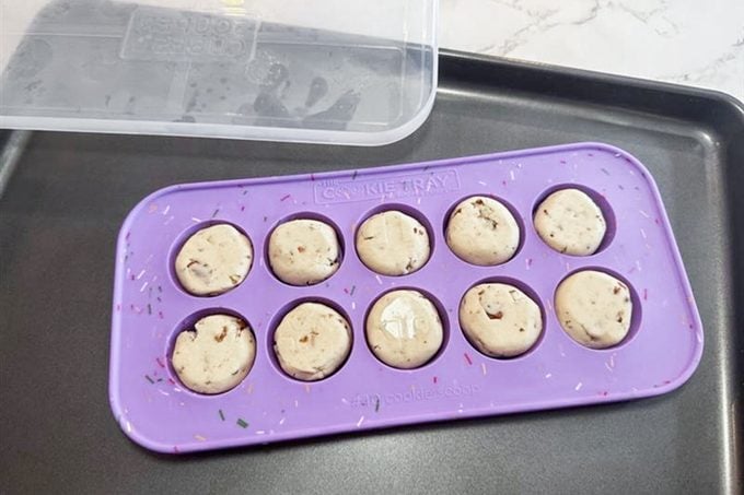 A purple silicone mold with ten round, unbaked cookie dough balls sits on a black baking tray. A clear plastic lid is partially visible at the top left corner.