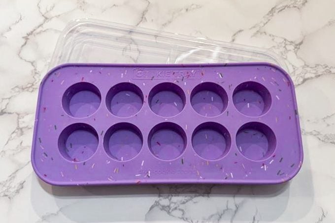 A purple silicone mold tray with ten round cavities and a clear lid, placed on a white marble surface.