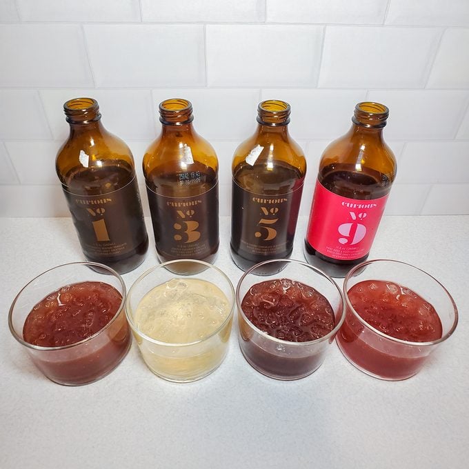 Four brown bottles with numbered labels stand behind four matching cups of colored jelly on a white countertop, against a white tiled background. The jellies correspond in color to the bottles behind them.
