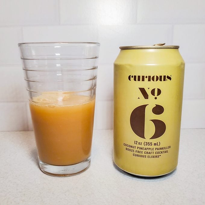 A glass filled with an orange-brown beverage sits next to a yellow can labeled "Curious No. 6" on a light countertop, with a white tiled wall in the background.