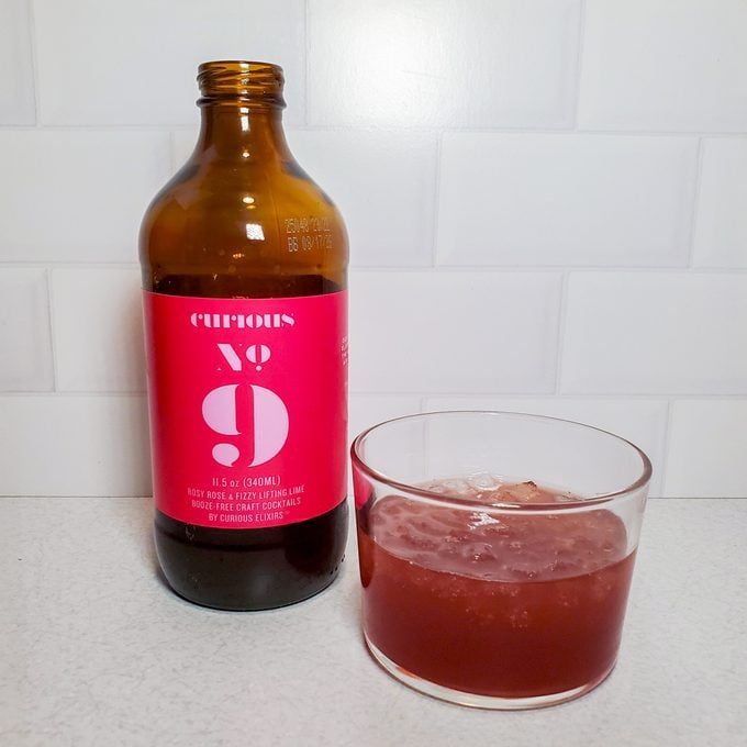 A brown glass bottle with a bright pink label reading "Curious No. 9" stands next to a clear glass filled with a reddish-brown beverage on a white countertop, against a white tiled background.